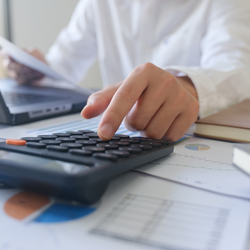 Comptabilité et Calcul : Main avec Calculatrice et Graphiques Gros plan sur un doigt appuyant sur une calculatrice noire posée sur des graphiques financiers, un homme en chemise blanche travaille sur un ordinateur portable.