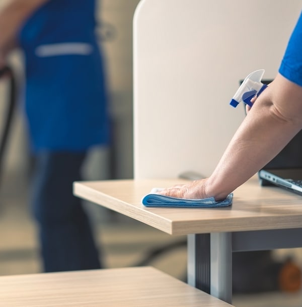 Nettoyage de bureau : désinfection et entretien ménager Main d'un agent d'entretien désinfectant un bureau avec un chiffon bleu et un vaporisateur, une autre personne est floue à l'arrière.
