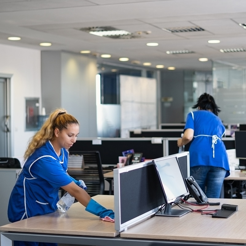 Nettoyage de bureau : femmes désinfectant des postes de travail Femme en uniforme bleu désinfectant un bureau avec un spray et un gant, une autre femme nettoie en arrière-plan dans un open space.