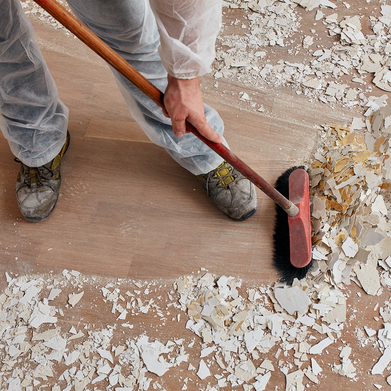 Nettoyage après démolition : balayer débris de plâtre Ouvrier en combinaison blanche balayant des débris de plâtre cassés sur un sol en bois lors de travaux de rénovation.