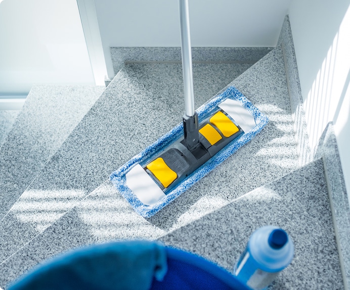 Nettoyage de marches en granit avec serpillière professionnelle Nettoyage d'un escalier en granit moucheté avec une serpillière microfibre bleue et jaune, lumière naturelle. Bouteille de produit visible.