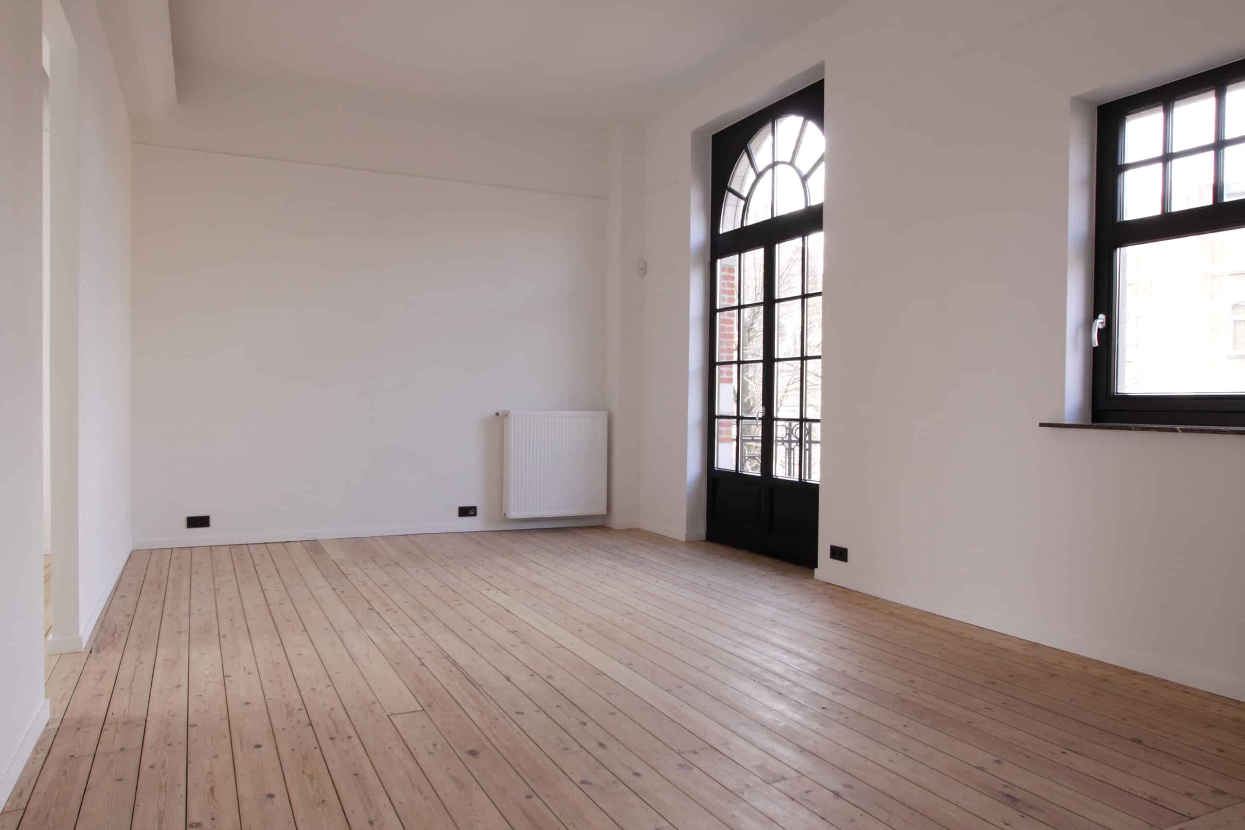 Pièce vide avec parquet clair, murs blancs, et une grande porte-fenêtre noire à arc en plein cintre donnant sur un balcon.